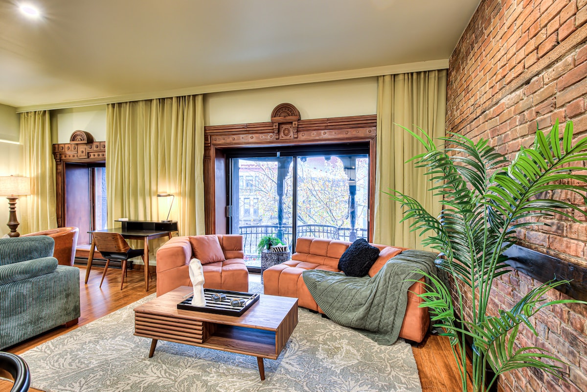 A spacious living area is highlighted by soft, warm lighting and large windows with elegant curtains. The room features a comfortable seating arrangement with a sectional sofa and accent chairs, complemented by a stylish coffee table and decorative plant, enhancing the overall inviting ambiance.