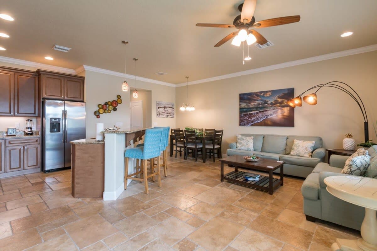 An open-plan living area features a fully equipped kitchen with wooden cabinets and stainless-steel appliances. A spacious dining table accommodates six, while comfortable seating arrangements invite relaxation. Soft lighting from decorative fixtures accentuates the light neutral walls and tiled flooring throughout the space.