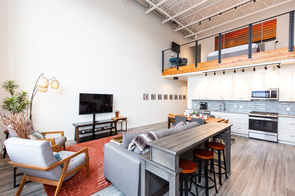 A modern living space features high ceilings, showcasing a loft area with a spacious sleeping arrangement. Comfortable seating is arranged around a large dining table. A kitchen with stainless steel appliances is visible, complemented by a sleek backsplash. Natural light enhances the open layout.