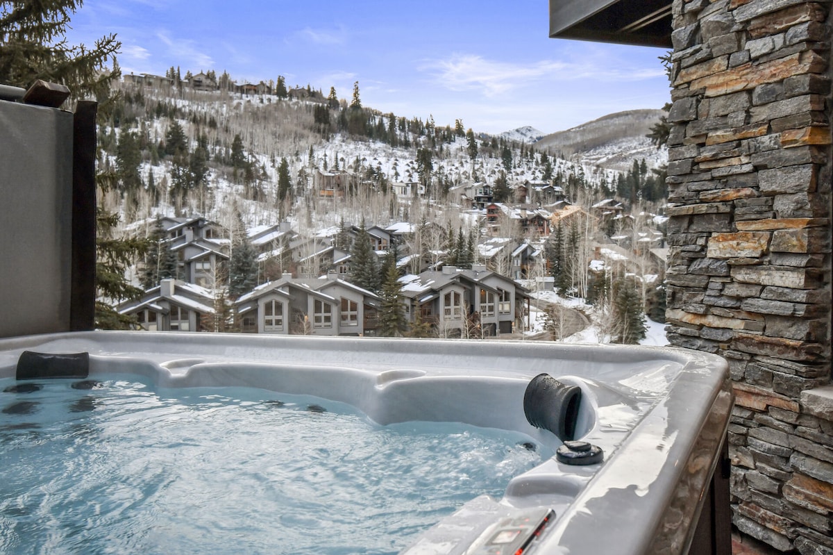 A hot tub surrounded by a stone wall offers a relaxing view of a snowy landscape. Rolling hills and mountain ranges are visible in the background, with quaint townhouses nestled within the trees, enhancing the sense of serenity.