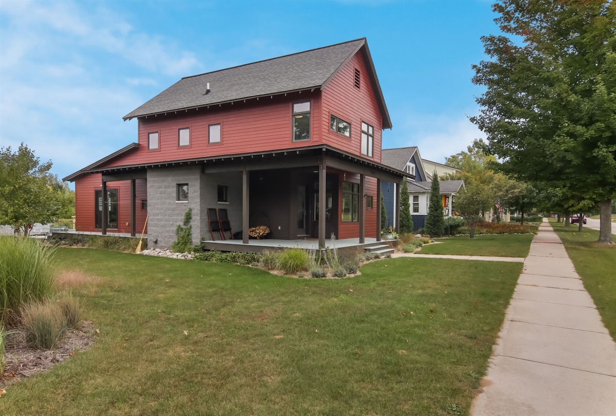 The exterior of Borealis Haus is shown, featuring a combination of red and gray siding. A spacious porch with seating is visible, accompanied by landscaped green grass and shrubs. The home is surrounded by trees and located on a peaceful, paved walkway.