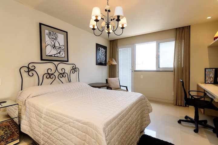 Slaapkamer met queensize bed, hoofdeinde, kussens, deken, kroonluchter, nachtkastje, lamp, tapijt, raam, jaloezie, gordijnen, tafel/bureau, bureaustoel, planken.