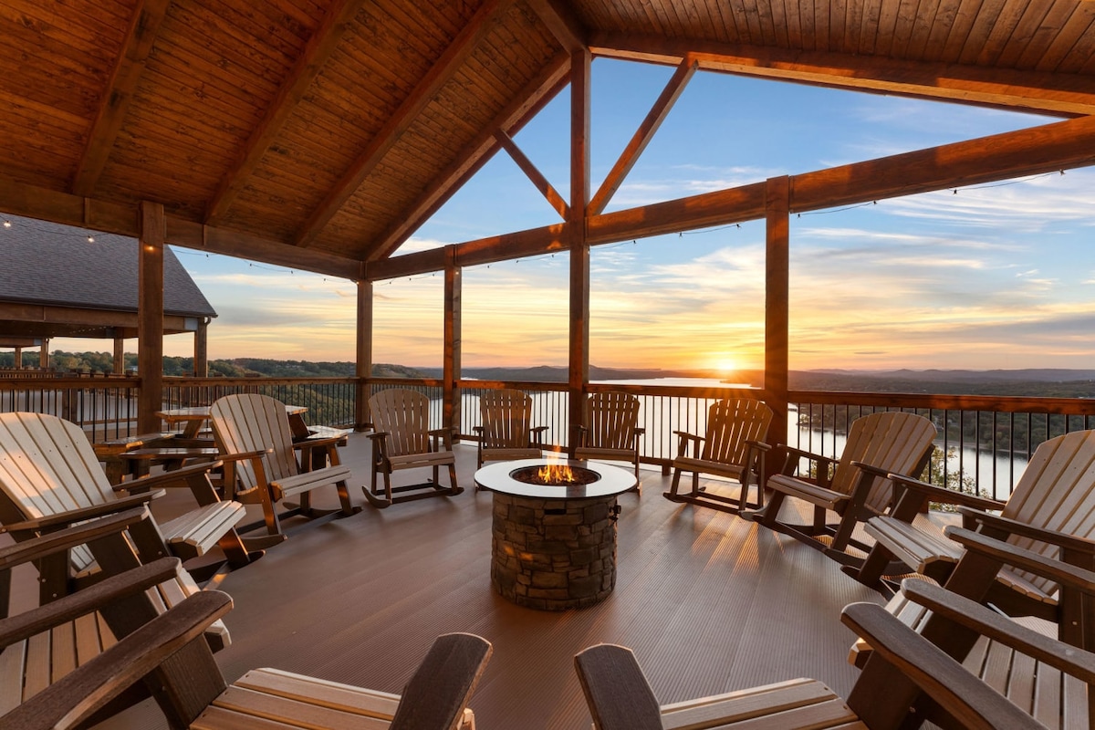 A spacious deck features multiple wooden Adirondack chairs arranged around a central stone fire pit. The backdrop showcases a stunning sunset over the lake, with warm colors reflecting on the water and a gently lit sky.