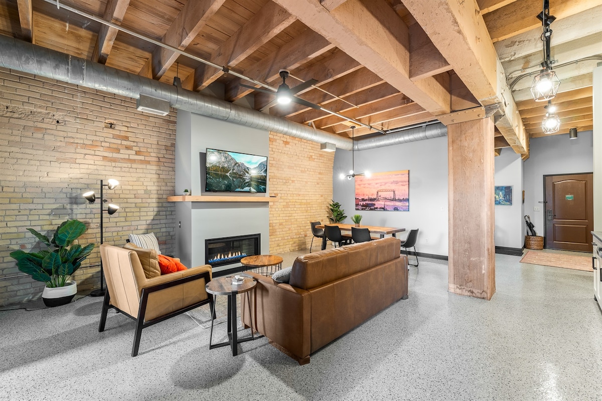 A spacious living area features a contemporary design with a mix of seating options, including a tan leather sofa and modern armchair. Exposed beams and brick walls add character, while artwork continues the theme of the surrounding scenery. A dining area with a wooden table is arranged nearby.
