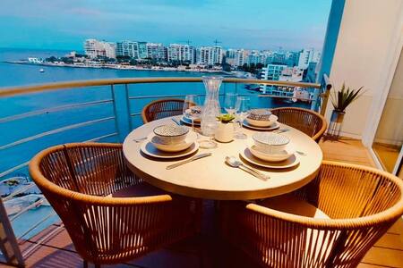 A round dining table is set with elegant dinnerware, featuring plates, glasses, and utensils for four. The table is surrounded by wicker chairs and overlooks the shimmering waters of Balluta Bay, with a backdrop of the coastal skyline, illuminated by soft evening light.