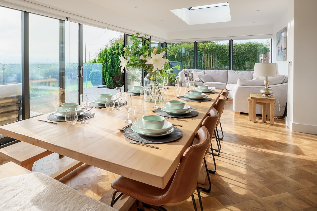 A modern dining area features a large wooden table surrounded by eight tan leather chairs. Elegant table settings with plates and glassware are arranged for a meal. Floor-to-ceiling windows allow natural light to illuminate the space, offering views of the lush outdoor surroundings.