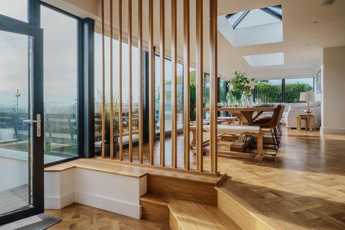 A light-filled open-plan space showcases stylish wooden flooring and large glass windows, offering views of the surrounding landscape. A wooden dining table can be seen, accompanied by an assortment of chairs. Elegant wooden accents and greenery enhance the modern aesthetic.
