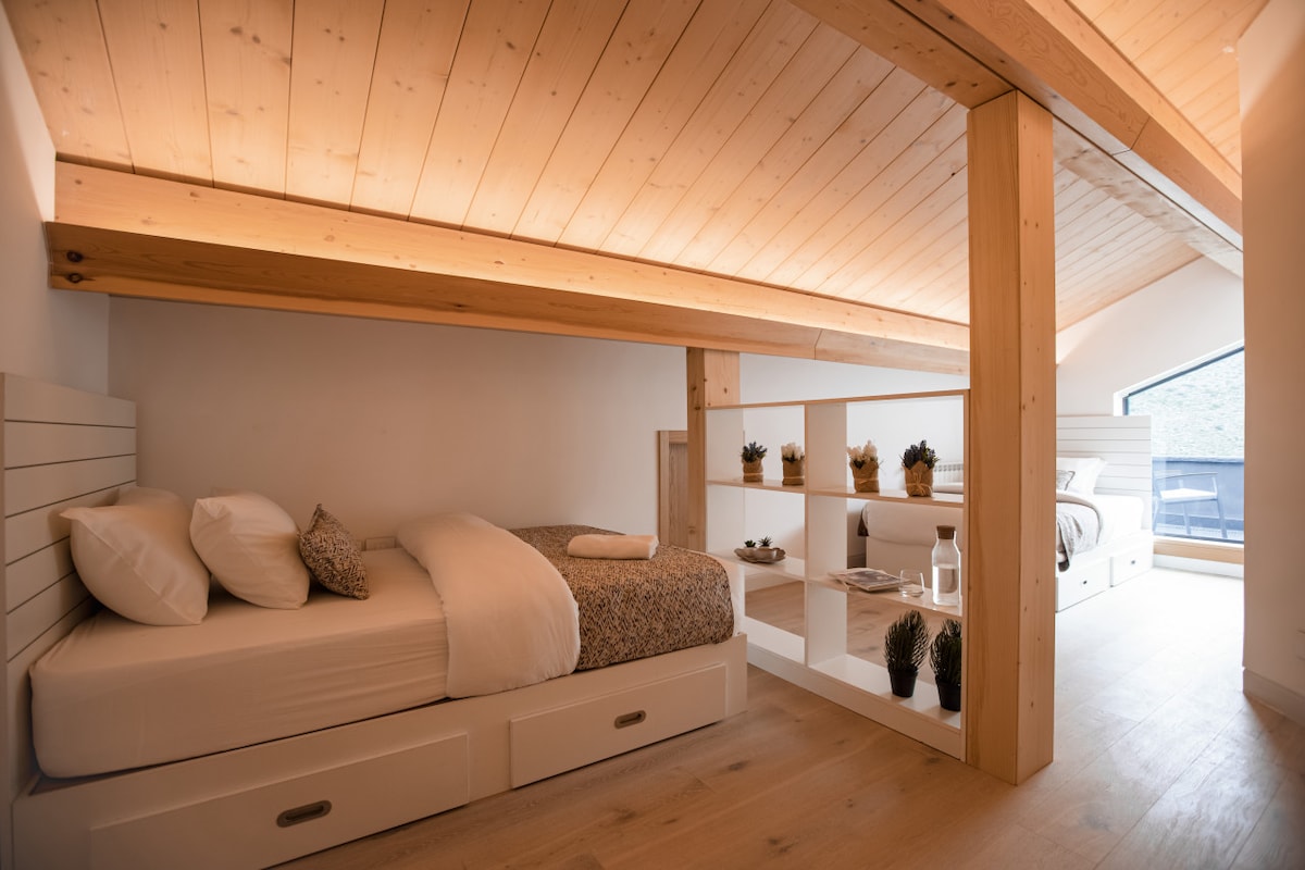 The image shows a cozy loft-style bedroom with sloped wooden ceilings. Two beds are positioned, with one featuring a patterned coverlet. A partition bookshelf with small plants is visible, and natural light enters through a window that leads to a balcony.