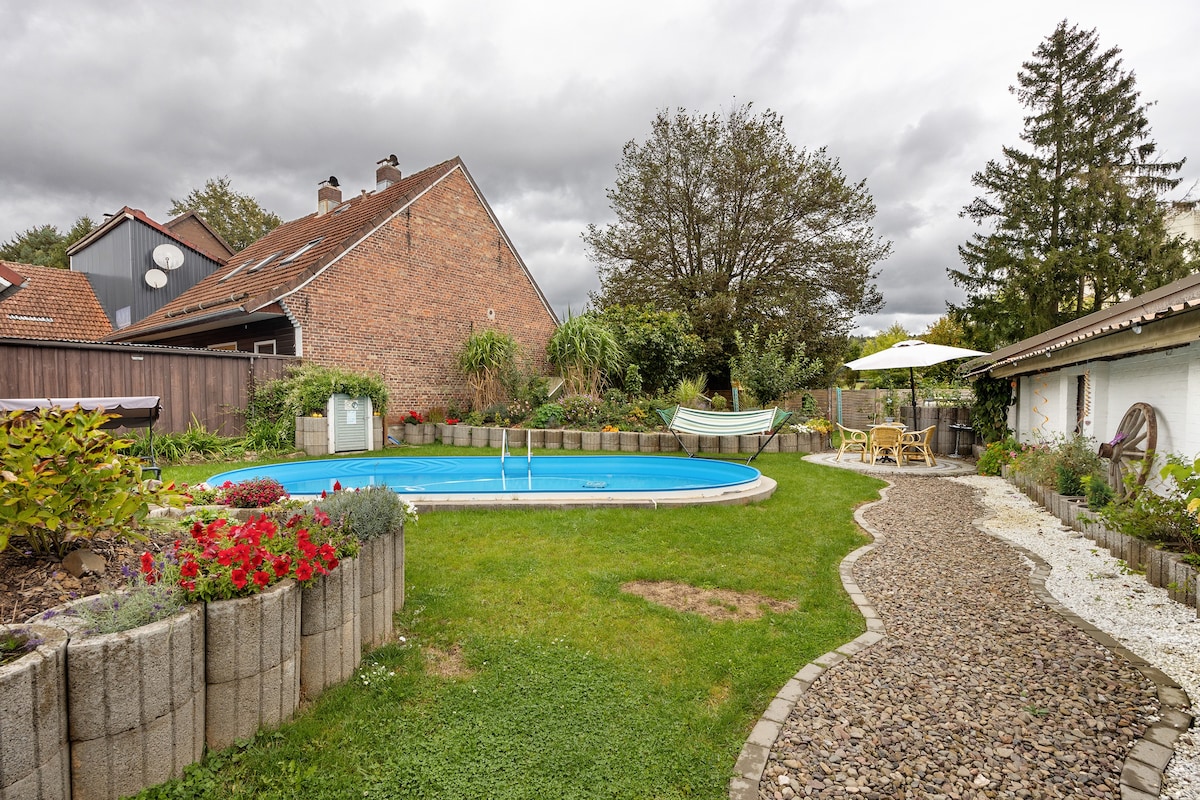 A spacious outdoor area features a circular pool surrounded by a well-maintained lawn and flower beds. A winding gravel pathway leads to seating arrangements under shaded umbrellas, while trees and hedges provide a natural backdrop.