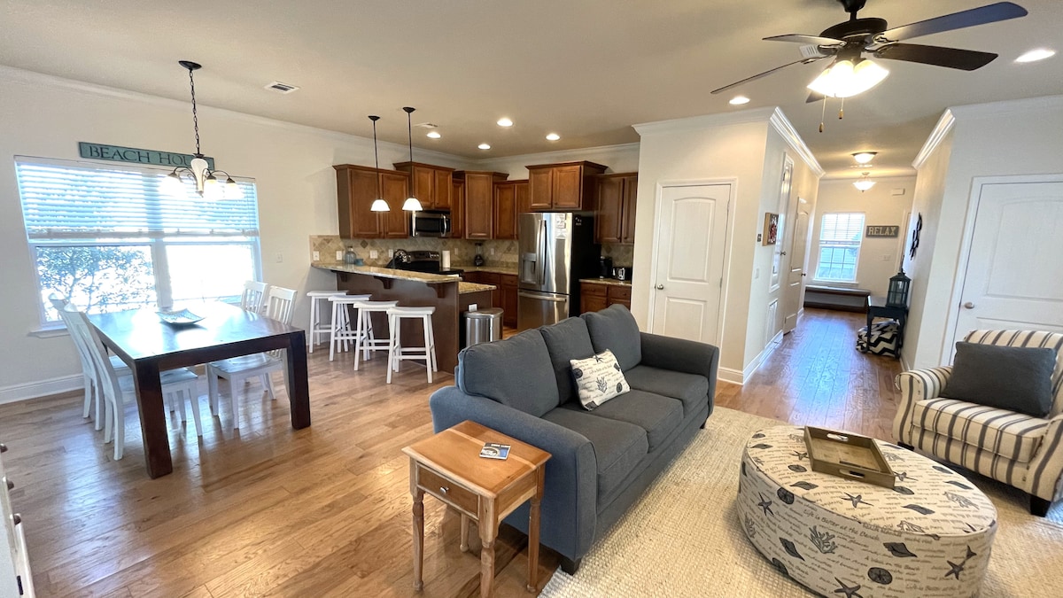 An open-concept living area is displayed, featuring a comfortable gray sofa, a patterned area rug, and a light-colored coffee table. A dining table with white chairs is positioned adjacent to a well-equipped kitchen with wooden cabinetry, modern appliances, and warm lighting.