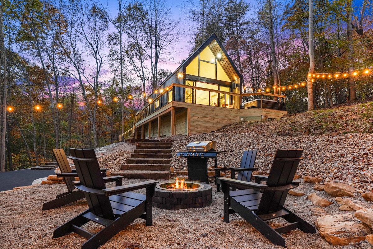 An outdoor space features a fire pit surrounded by four black wooden chairs, providing a cozy gathering area. Behind, a modern cabin is framed by trees, with large windows reflecting the evening sky and string lights illuminating the area.