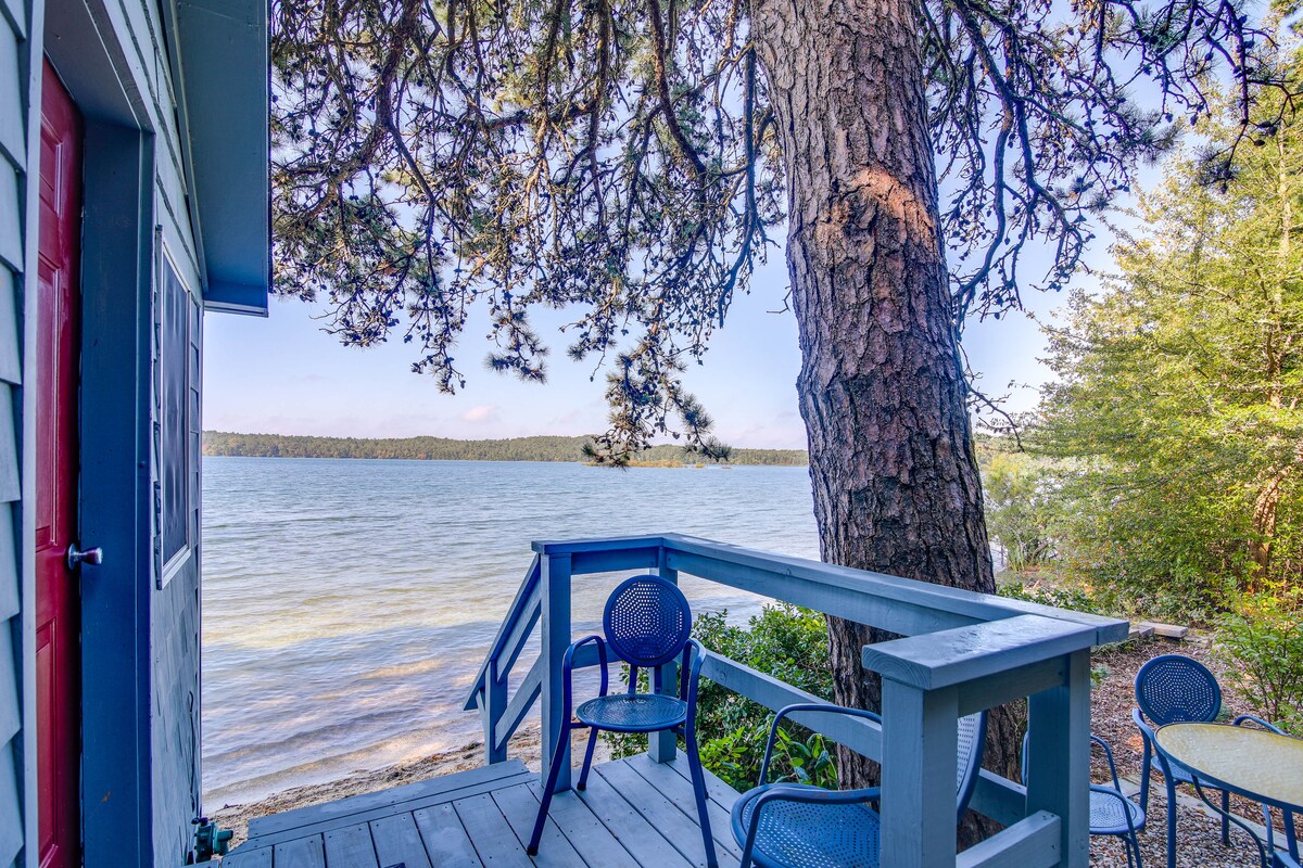 A furnished deck overlooks Great South Pond, featuring a table and two chairs positioned under the shade of a large tree. The serene water and surrounding greenery can be seen in the background, creating a peaceful outdoor setting.