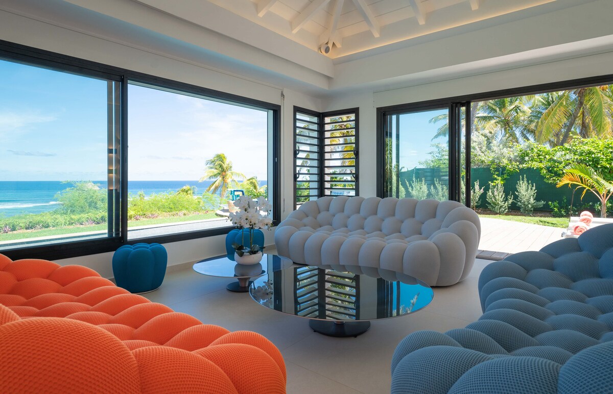 A spacious living area features contemporary seating in vibrant hues of orange and blue, arranged around a sleek round table. Expansive windows frame a stunning sea view, inviting natural light and providing a connection to the outdoor landscape.