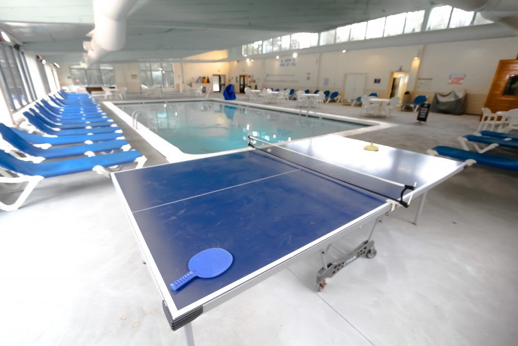 An indoor recreational area features a blue ping pong table positioned near a spacious saltwater pool. Lounge chairs line one side, providing seating for relaxation. Bright overhead lighting enhances the airy ambiance, while large windows allow natural light to fill the space.