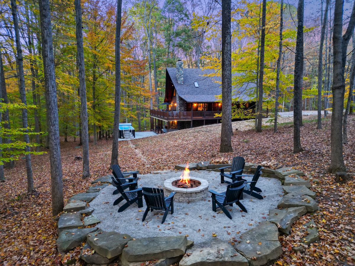 An outdoor fire pit area is surrounded by stone seating, with a crackling fire resting in the center. Towering trees with autumn foliage create a serene backdrop, while the log home stands illuminated in the distance. Warm colors enhance the tranquil ambiance of the setting.