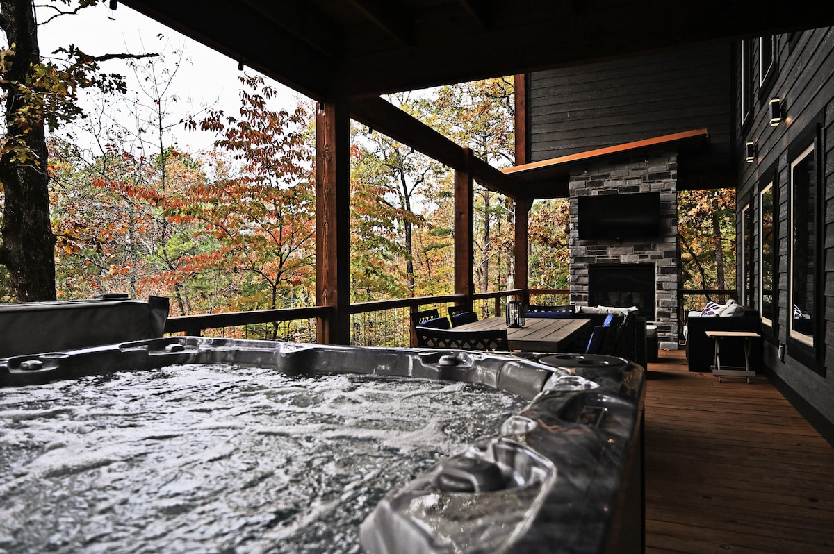 A hot tub is situated on a spacious deck, surrounded by wooden beams and a scenic view of autumn foliage. A cozy outdoor fireplace is visible nearby, complementing the serene atmosphere offered by the nature that envelops the area.