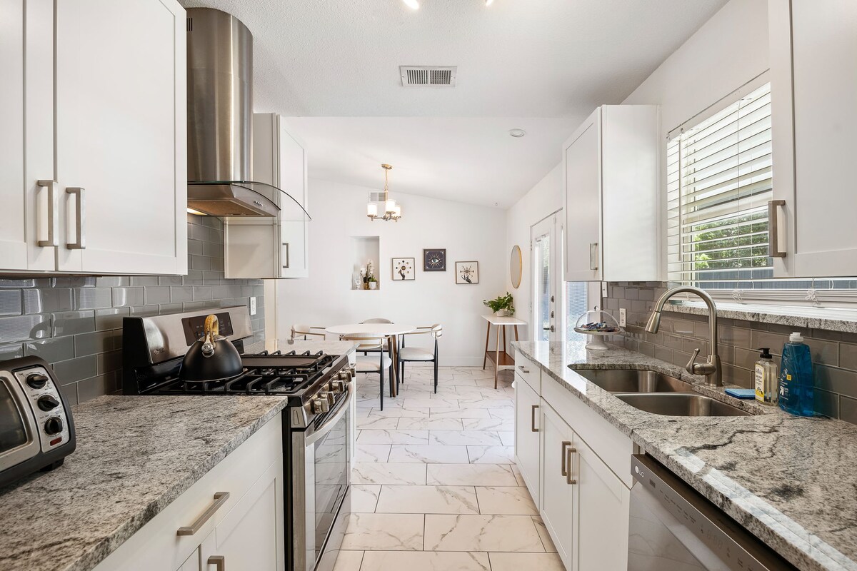A modern kitchen area features white cabinetry and sleek granite countertops. Stainless steel appliances are visible, including a gas stove and microwave. Natural light fills the space through windows, while a dining area is glimpsed in the background with a small table and plants.