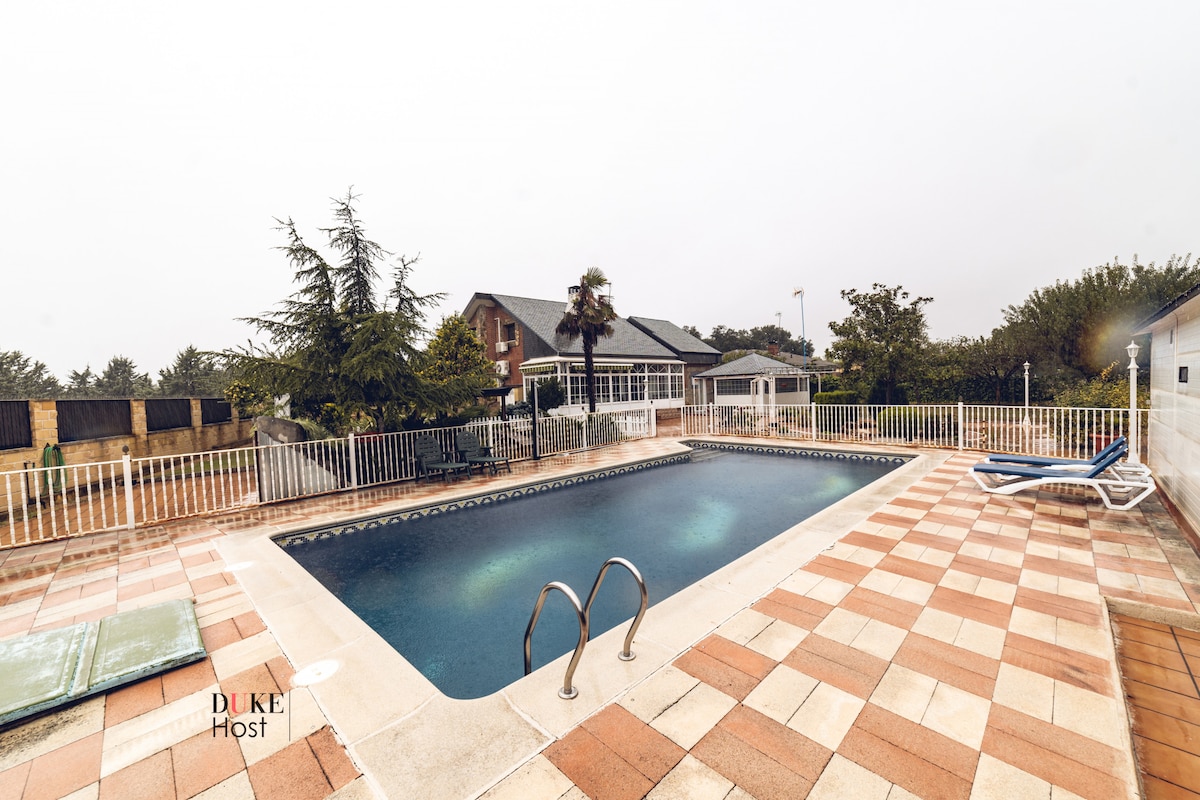 A private swimming pool is set within a fenced area, surrounded by tiled decking. Lush trees and greenery can be seen in the background, while a covered seating area is visible at one end of the pool. The atmosphere is calm and serene.
