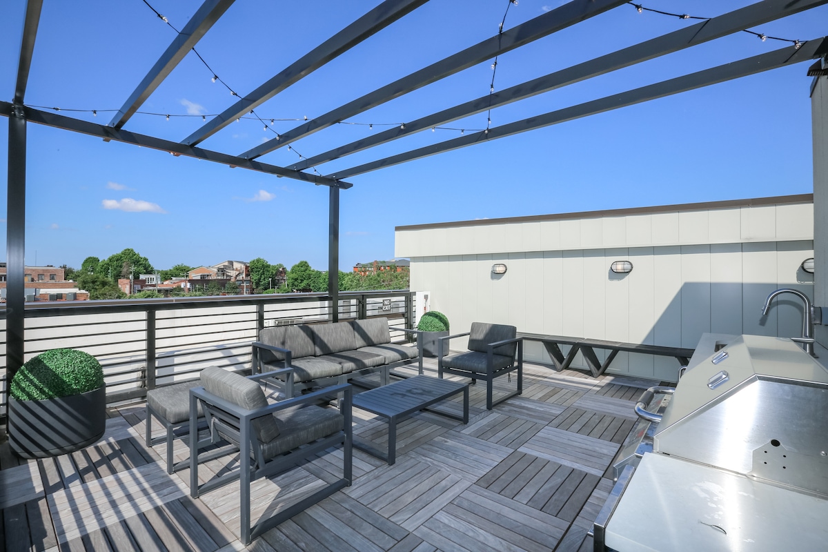 A rooftop lounge is shown, featuring comfortable seating arranged on a wooden deck. A grill is positioned nearby, with planters adding greenery to the space. The surrounding area displays urban views under a clear blue sky.