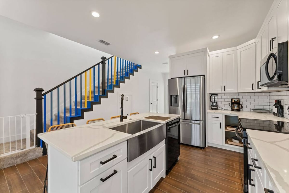 A modern kitchen is showcased, featuring white cabinetry and stainless steel appliances, including a refrigerator and microwave. A large island with a farmhouse sink is complemented by stylish seating. A vibrant staircase with blue and yellow accents stands in the background, enhancing the open layout.