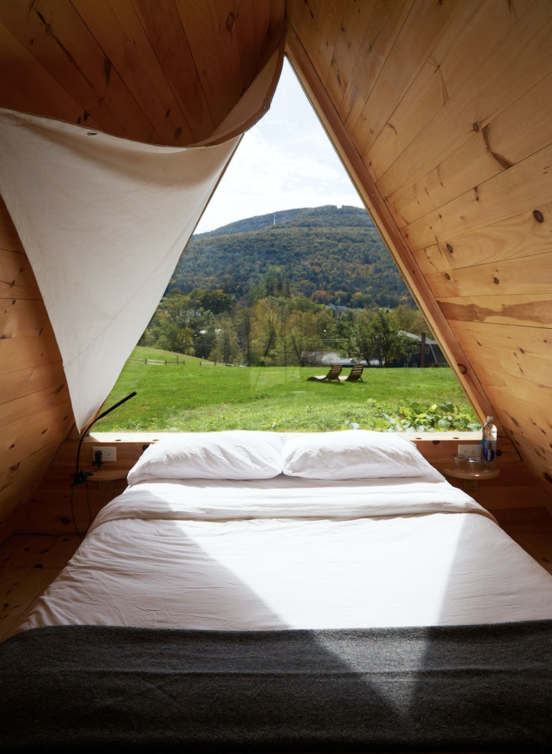 A cozy sleeping area is framed by wooden walls, featuring a spacious bed dressed in crisp white linens. A triangular window offers a view of rolling green hills and outdoor seating, allowing natural light to fill the room.