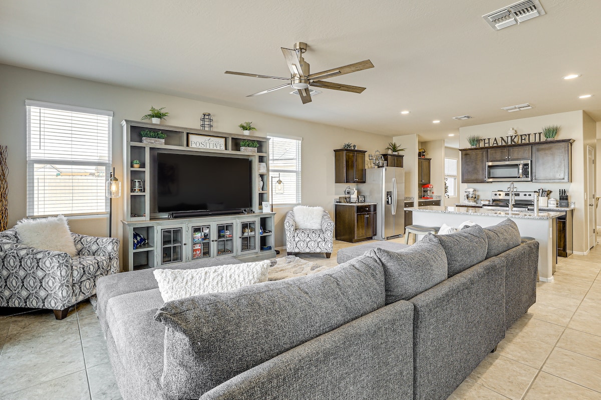 A modern living area is shown with a spacious grey sectional sofa facing a large flat-screen TV. Light-colored cabinets and countertops highlight the open kitchen in the background. Natural light enters through multiple windows, complemented by decorative plants on shelves and counters.