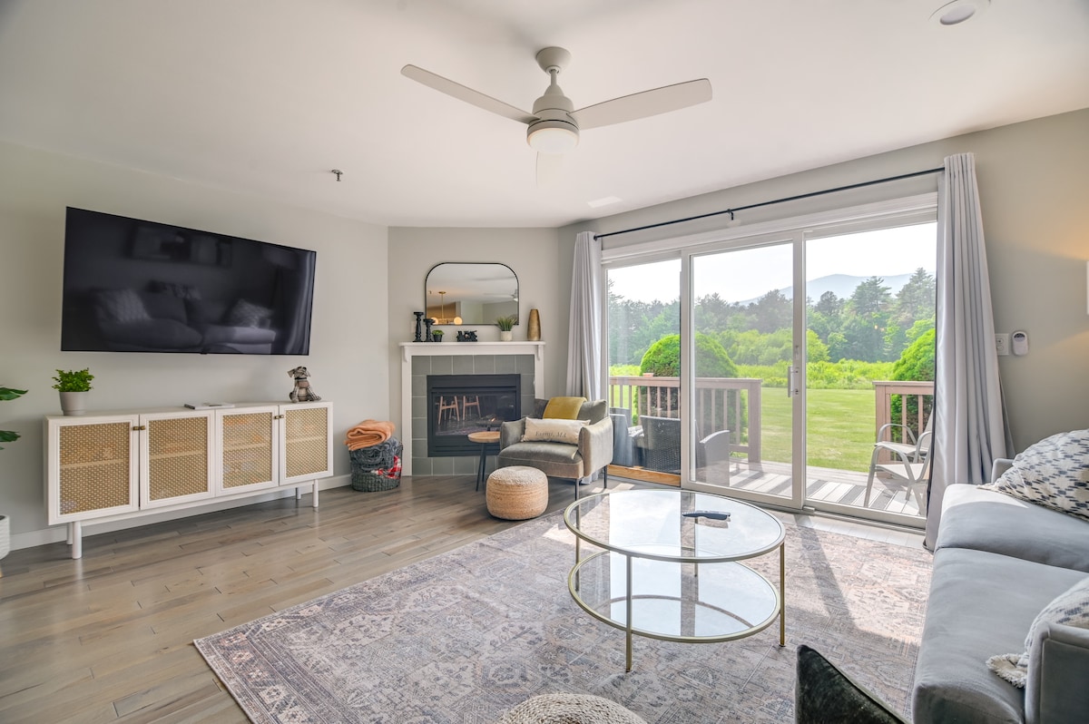 A welcoming living area includes a large flat-screen TV mounted on the wall. A glass coffee table sits on a patterned rug, and seating is provided by a light-colored sofa. Natural light streams in through sliding glass doors that lead to a furnished deck and view of greenery.
