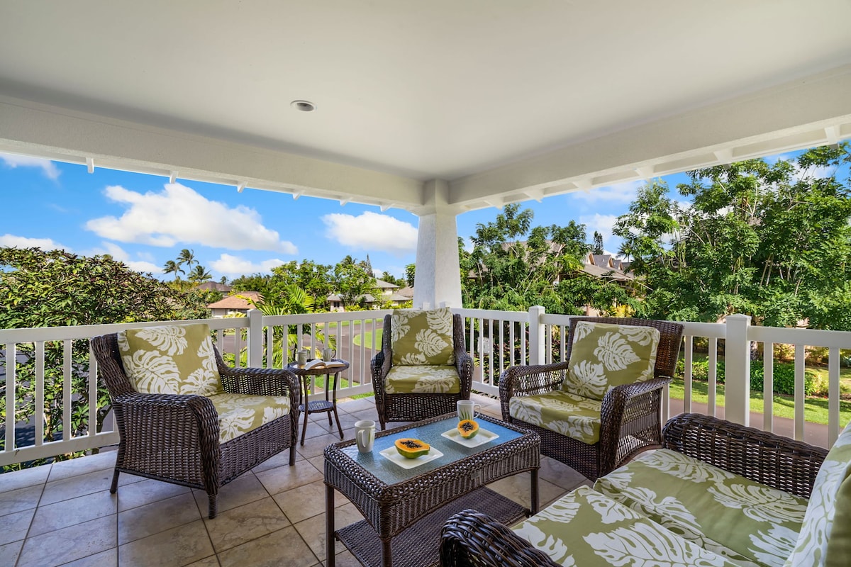 A covered lanai features comfortable seating with tropical-patterned cushions. A small coffee table is set between the chairs, and views of lush greenery and a clear sky are visible beyond the railing.
