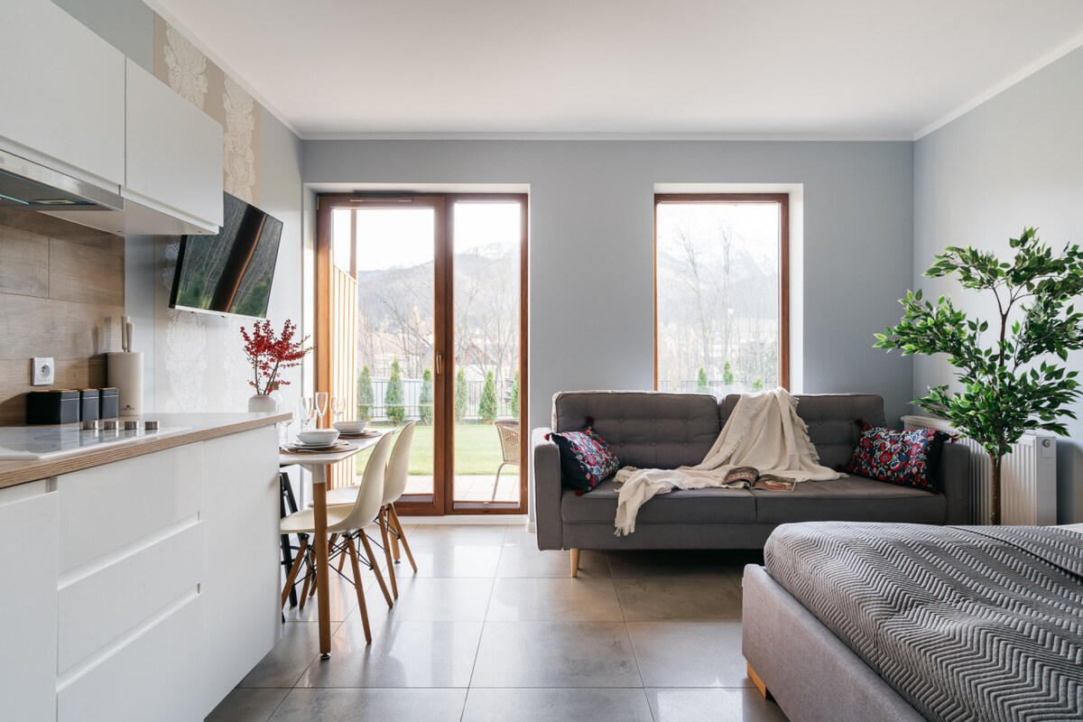A modern living area showcases a comfortable gray sofa, accompanied by a light blanket. A dining table with four chairs sits adjacent to the kitchen. Floor-to-ceiling windows provide views of the outdoor space and mountain landscape, allowing natural light to flow into the room.
