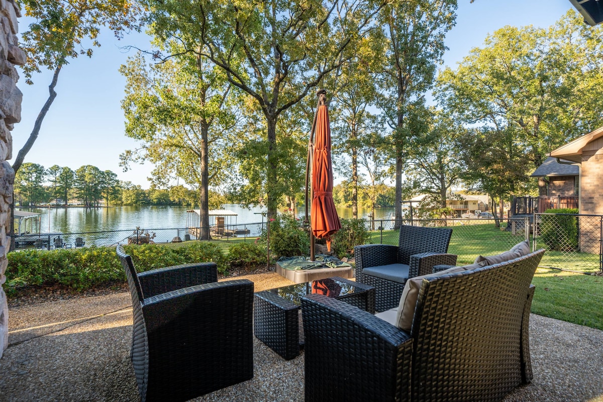 An outdoor seating area features comfortable wicker chairs arranged around a small coffee table beneath a large orange umbrella. Lush greenery surrounds the space, and the serene lake is visible in the background, framed by tall trees.