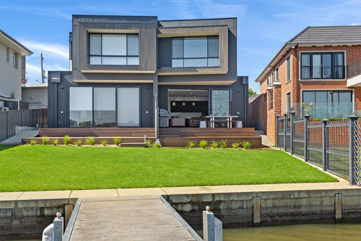 A modern two-story home is depicted, featuring a sleek exterior with large glass windows. The landscaped yard is complemented by manicured grass and a wooden deck, leading to a calm waterfront area.