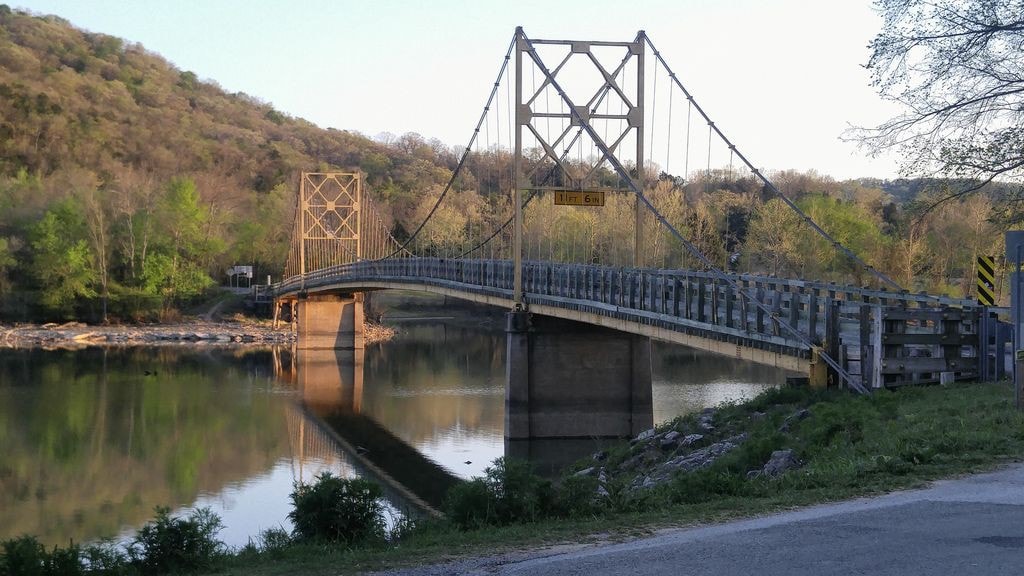 A suspension bridge spans over calm waters, reflecting its structure and nearby greenery. The landscape features rolling hills in the background and a shoreline lined with trees and shrubs, creating a serene natural environment.