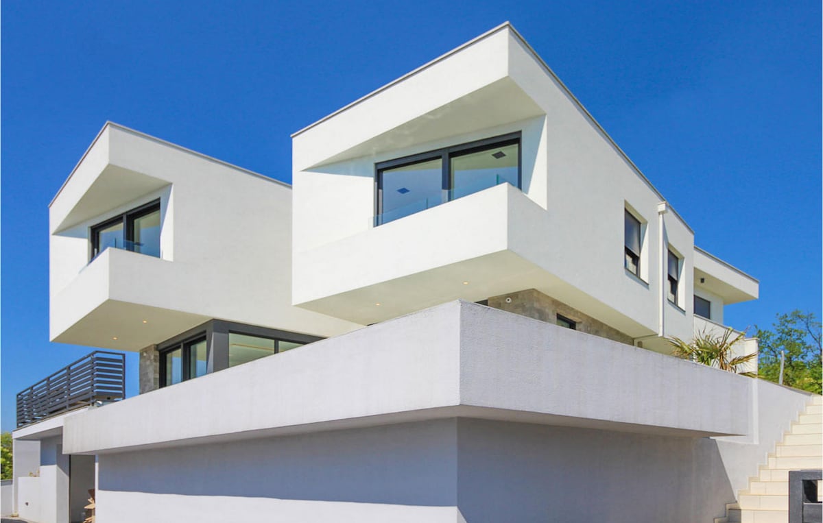 The modern villa features a unique architectural design with angular white facades and numerous large windows. Natural light floods the interiors, while the two-story structure is set against a clear blue sky, enhancing its contemporary appeal.