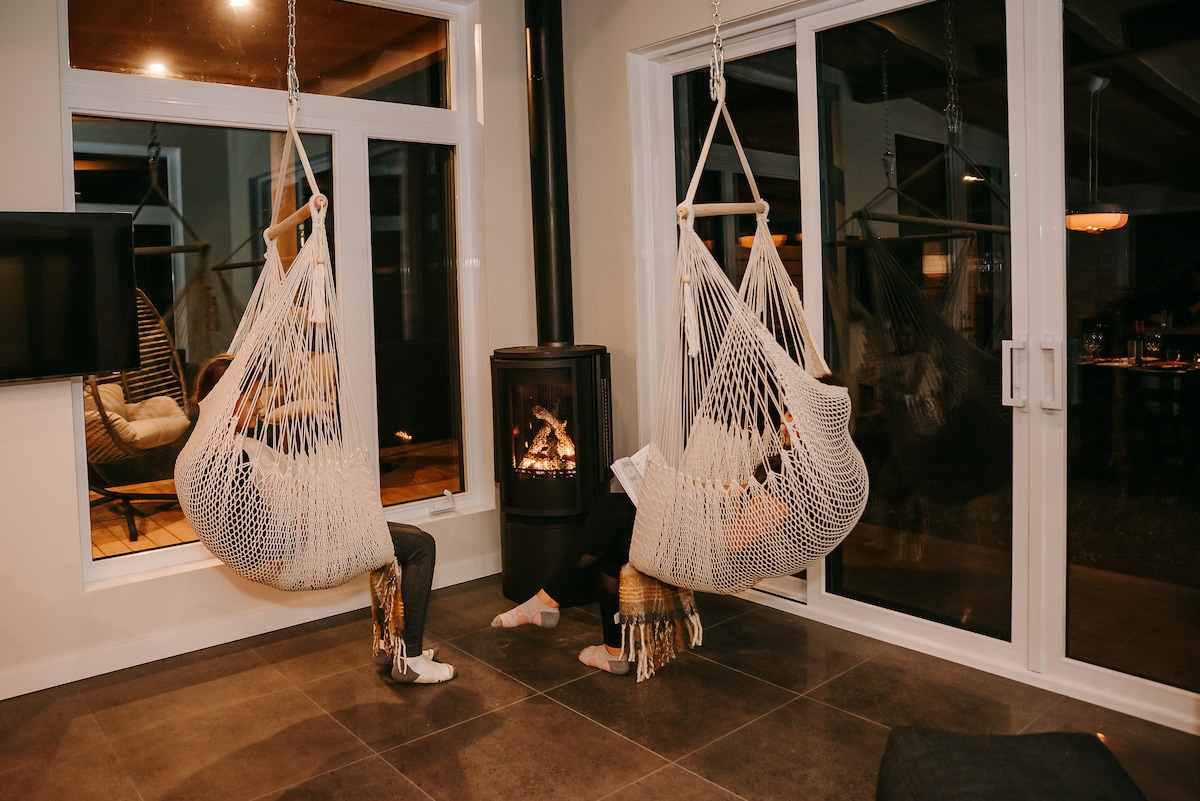 Two hanging chairs provide a cozy seating area near a modern fireplace. Large windows allow natural light to fill the space, offering views of the outdoor area. The floor is tiled, contributing to a clean and contemporary aesthetic.
