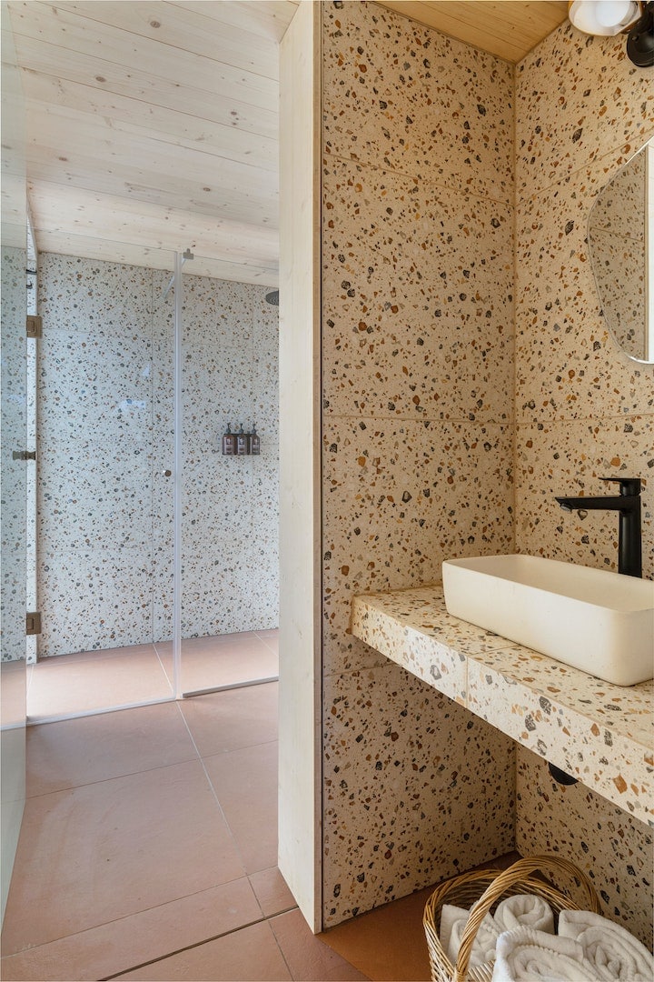 A modern bathroom features a textured stone wall and a spacious shower area with glass doors. A sleek sink is positioned on a light-colored countertop, complemented by a mirror above. Natural light fills the space, highlighting the warm tones of the flooring.