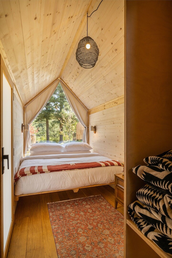A bright sleeping area features a spacious bed dressed in white linens with a red and white striped blanket. Natural light filters in through large windows, illuminating wooden walls and a cozy rug on the floor. Soft towels are neatly arranged on a shelf nearby.