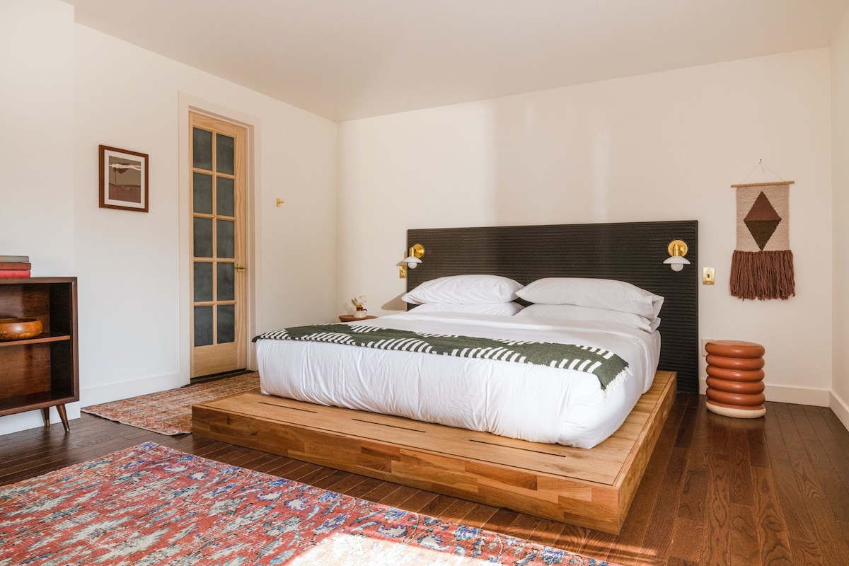 A modern bedroom features a spacious bed with neatly arranged white linens and a patterned throw. Soft lighting is provided by wall-mounted sconces. A wooden platform supports the bed, and a stylish area rug adds warmth to the hardwood floor.