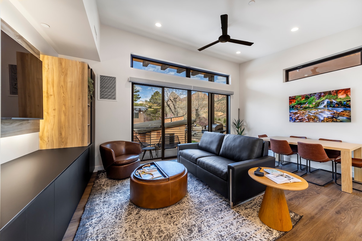 A modern living area features a leather sofa and a unique coffee table, with a Smart TV mounted on the wall. A large window offers scenic views of the surrounding landscape, while a dining table with several chairs is situated nearby, enhancing the inviting atmosphere.