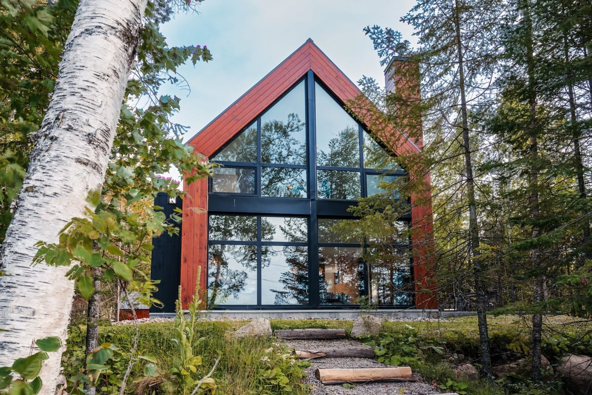The chalet features an A-frame design, with large glass windows reflecting the surrounding trees. A stone path leads from the greenery to the entrance, which is framed by vibrant foliage, inviting guests to explore the natural beauty of the location.