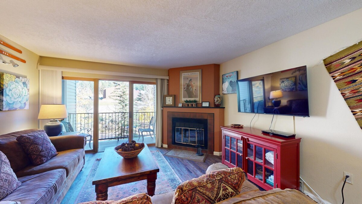 An open-concept living area features comfortable seating on a brown sofa and a patterned armchair. Natural light floods the space through large sliding glass doors leading to a private balcony. A flat-screen TV is mounted on the wall, and a decorative fireplace adds a cozy touch.