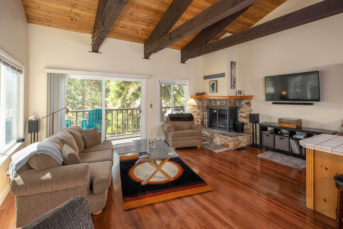 The living area features a spacious layout with wooden beams overhead and large windows that allow natural light to fill the room. Comfortable seating is arranged around a stone fireplace, accompanied by a flat-screen TV and a decorative area rug with a circular pattern.