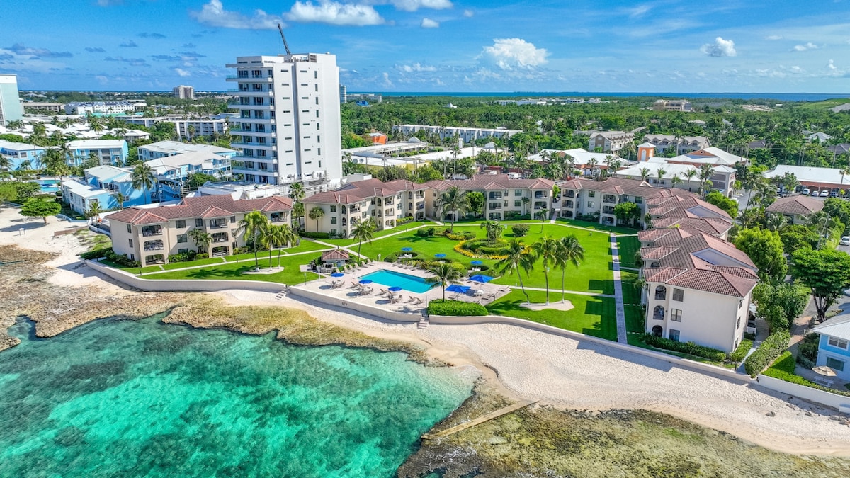 An aerial view highlights the oceanfront property, showcasing multiple two-story buildings surrounded by lush green lawns and palm trees. A swimming pool is visible adjacent to the sandy shoreline, which meets the clear turquoise waters, enhancing the inviting atmosphere of the complex.