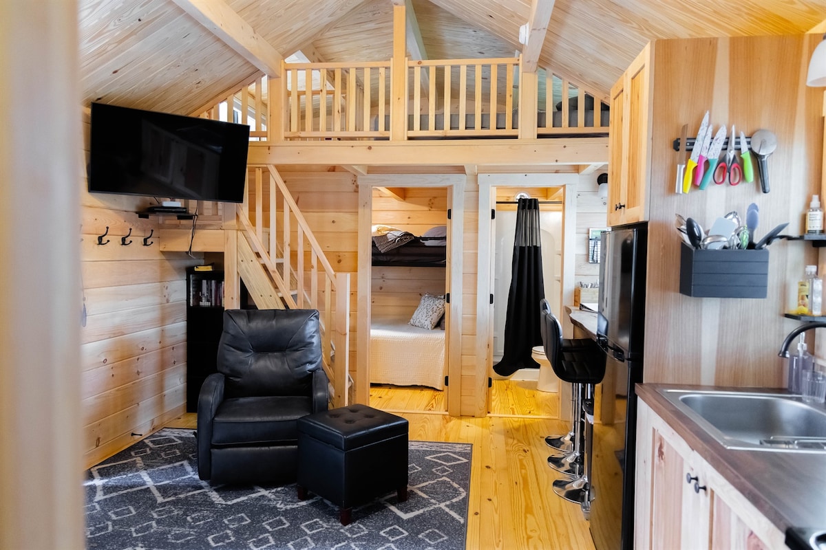 An inviting cabin interior features wood-paneled walls and ceilings, adding a warm touch. A comfortable black armchair sits in a cozy corner next to an ottoman. The fully equipped kitchen is arranged alongside the dining area, with bar stools positioned at a counter. A sleeping loft is visible above.