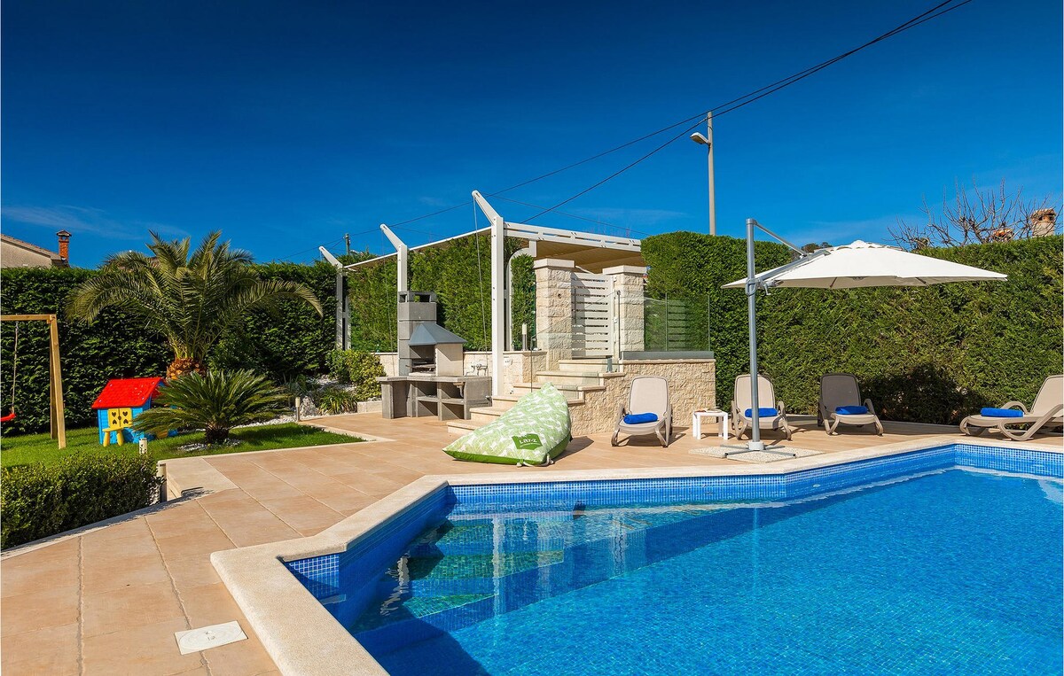 A vibrant outdoor area features a refreshing pool surrounded by comfortable sun loungers. A charcoal barbecue is set near a covered terrace, with a small play area visible. Lush greenery and bright blue skies provide a serene backdrop.