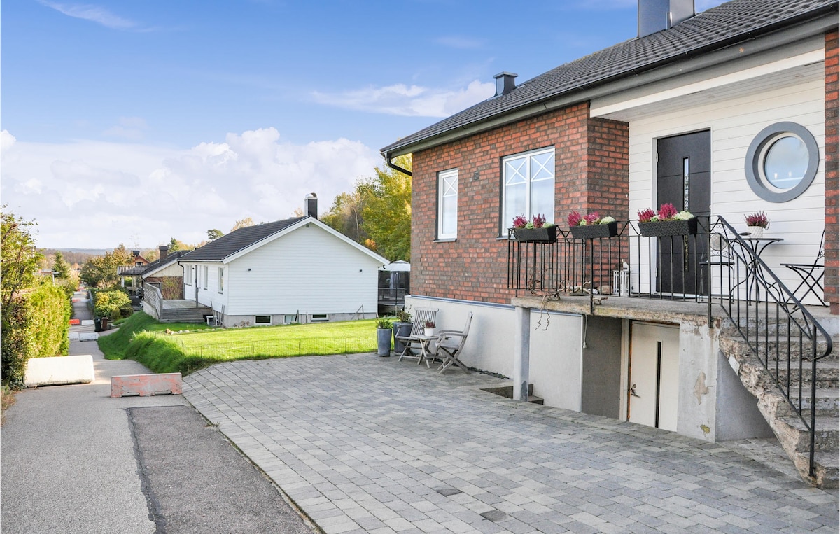 The exterior of the apartment features a contemporary brick façade with large windows and a circular window above the entrance. A set of stairs leads to the main door, while a paved path and garden area extend alongside the property, offering a view of neighboring houses.