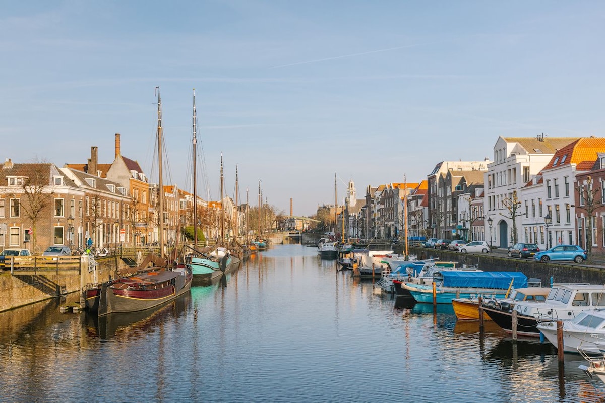 A tranquil canal scene showcases a collection of boats moored along the water's edge, surrounded by charming buildings. The clear sky reflects on the surface of the water, enhancing the serene atmosphere of the picturesque harborside location.