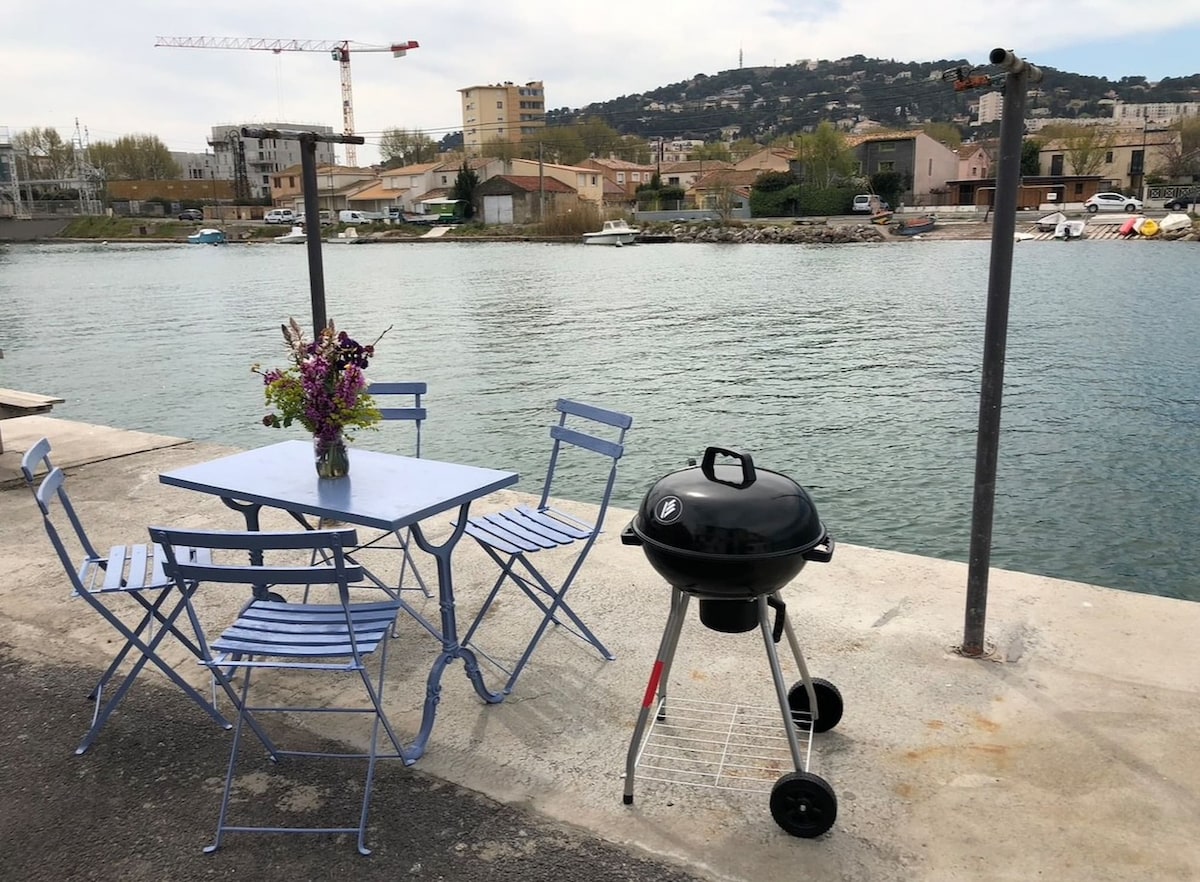 A small outdoor dining area is positioned by the water's edge, featuring a blue table and two matching chairs. A charcoal grill stands nearby, with a vase of fresh flowers adding a touch of color. The calm water reflects the surrounding buildings and greenery.