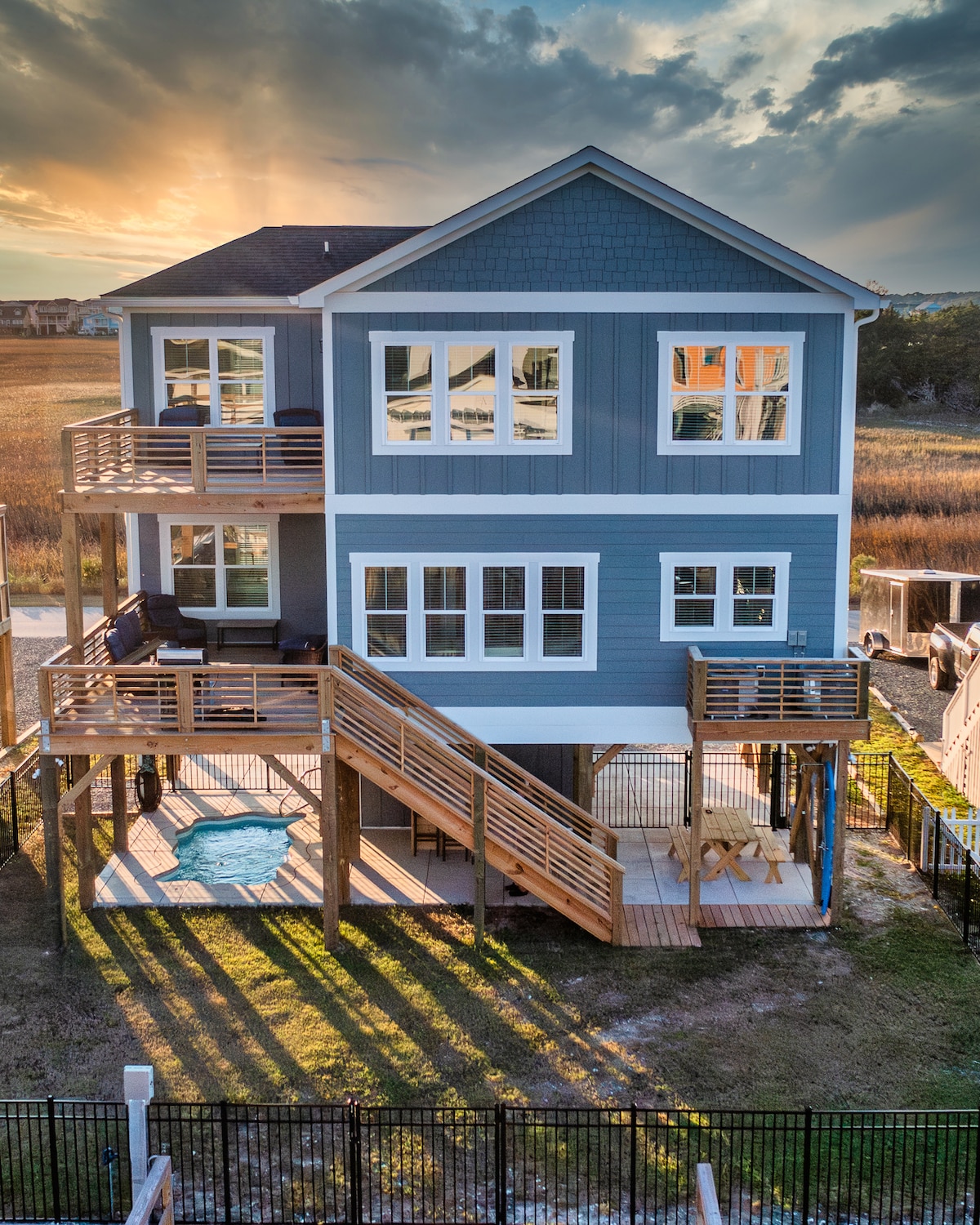 The exterior of a two-story blue house features multiple large windows reflecting the sunset. A wooden deck provides outdoor seating, with a set of stairs leading down to a yard. Shadow patterns are created on the grass by the setting sun.