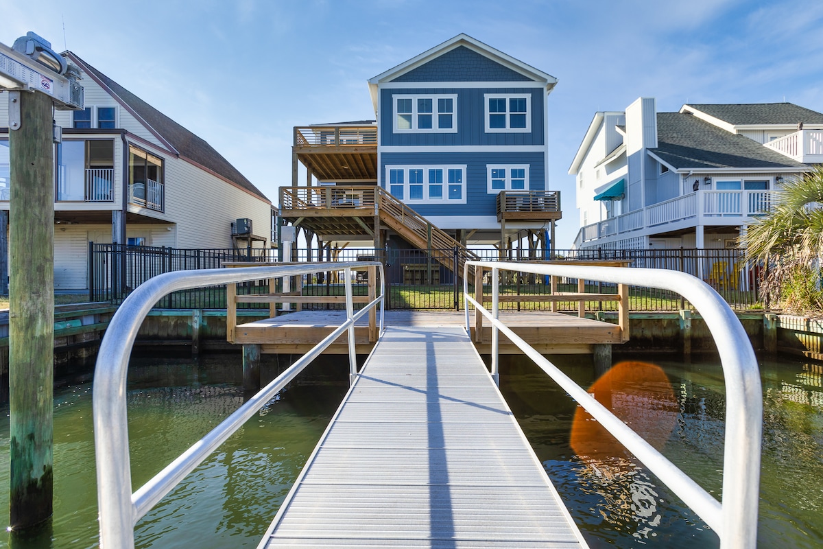 A long, sturdy dock extends over calm water, leading to a two-story blue house with deck access. The house features multiple outdoor spaces and is surrounded by fencing, with additional properties visible on either side. Clear skies are present overhead.