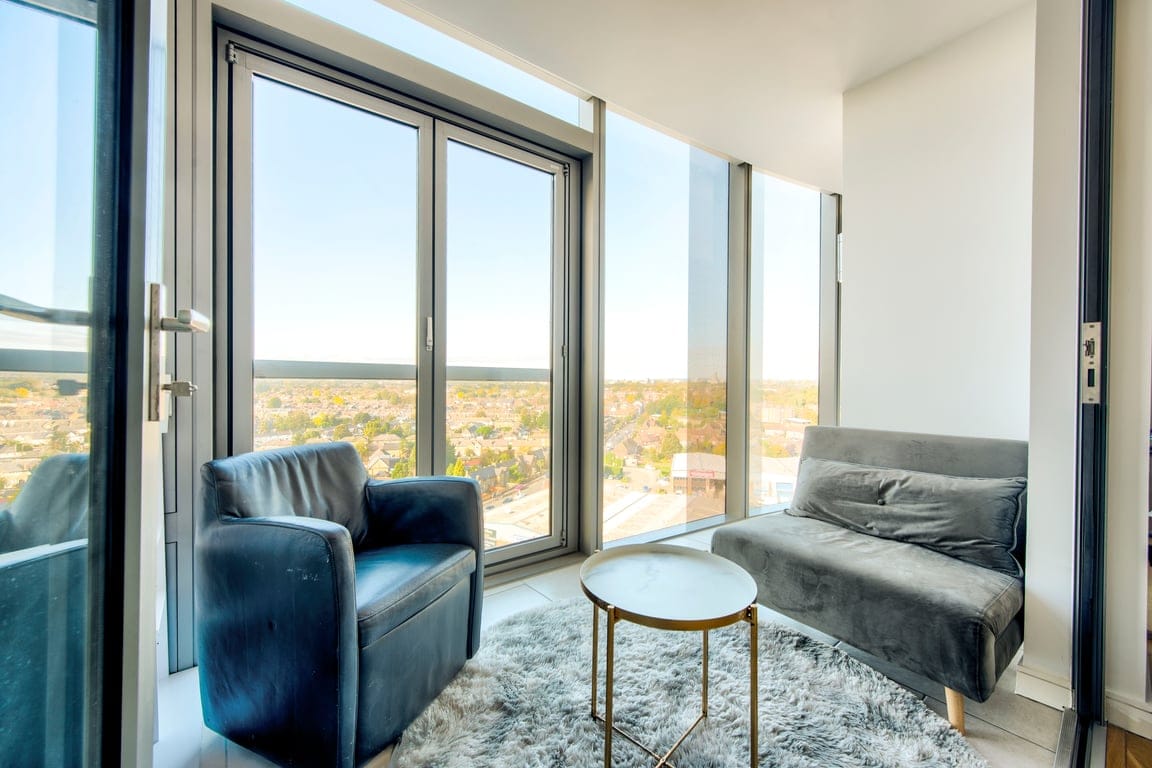 A cozy living area is presented with floor-to-ceiling windows offering expansive views of the surrounding landscape. A sleek round table sits between a velvet sofa and an upholstered armchair, complemented by a soft area rug beneath.